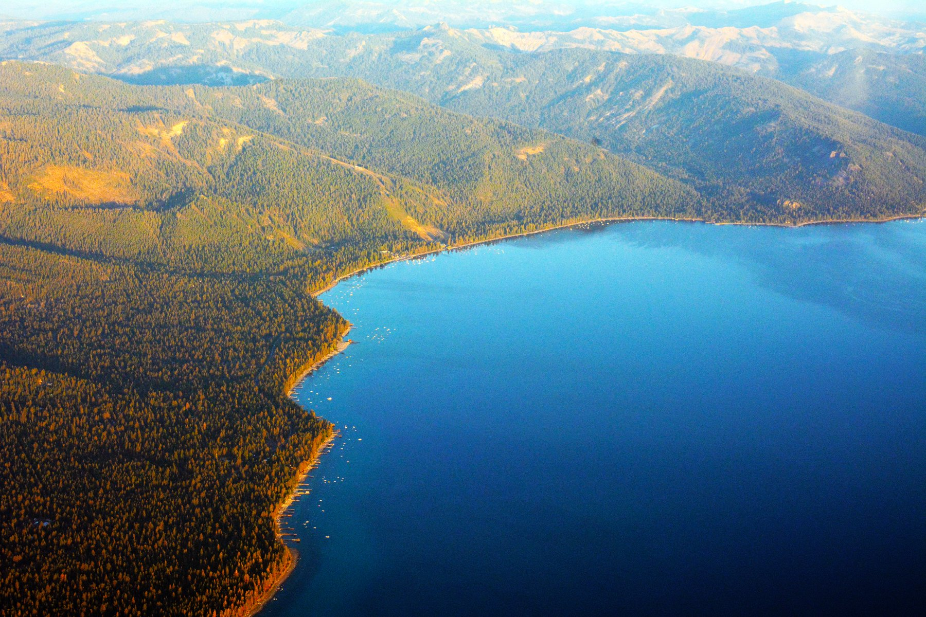 Aerial view of Lake Tahoe's forested shoreline and deep blue water with the Sierra Nevada mountains in the background.